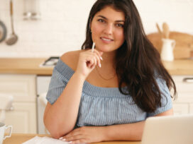 Smiling woman sitting at a table with a laptop, documents, and coffee, with a modular kitchen in the background.