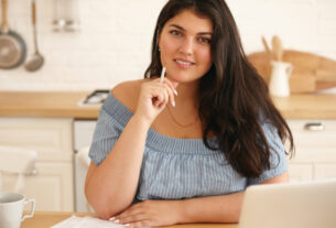 Smiling woman sitting at a table with a laptop, documents, and coffee, with a modular kitchen in the background.